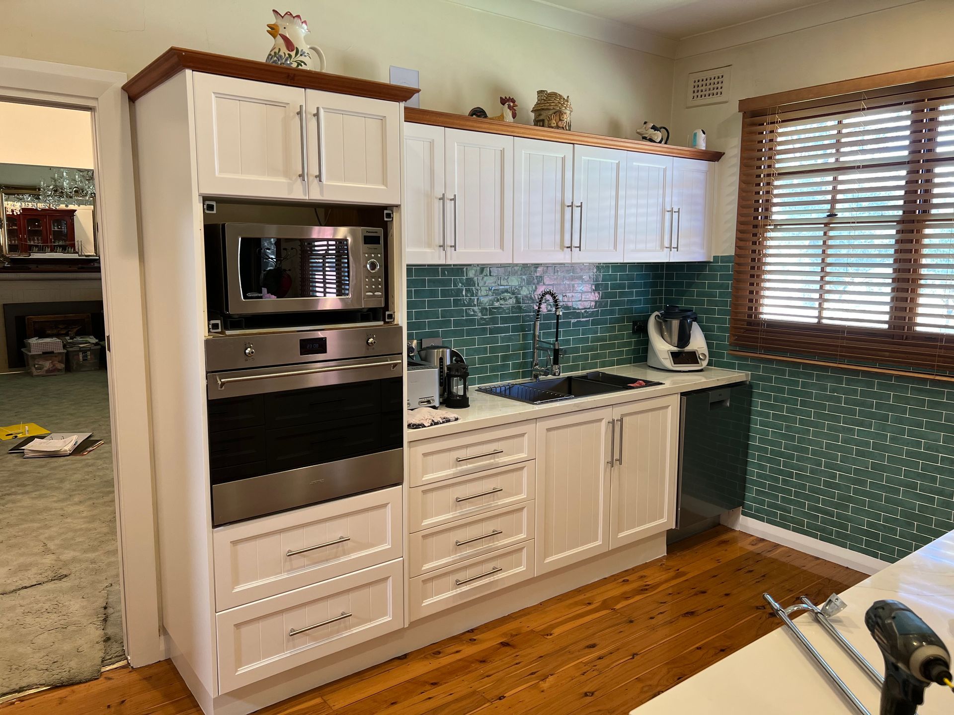 White kitchen with a built-in microwave and oven, green backsplash, and a wooden window.