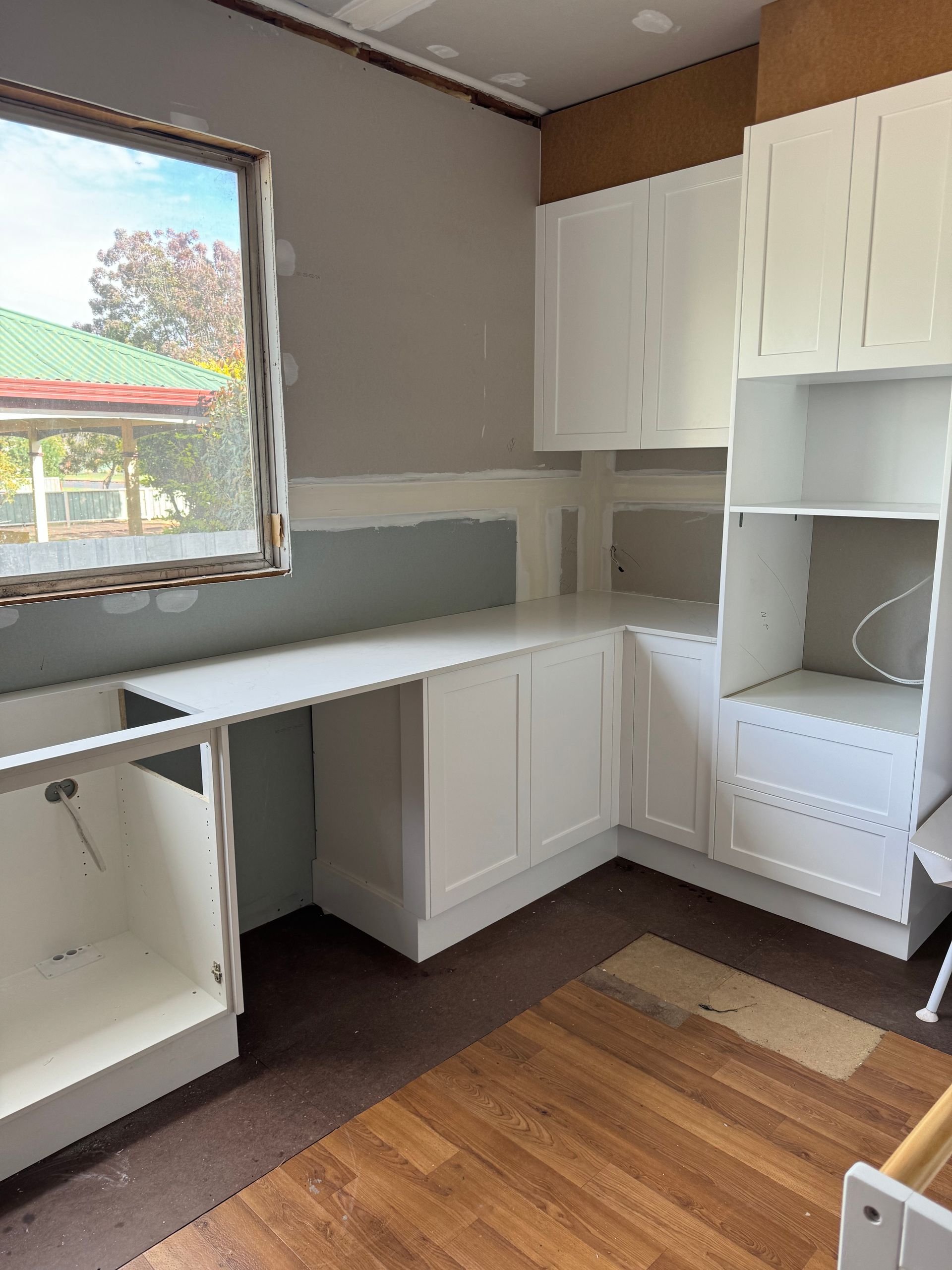 Interior view of a kitchen under construction, showing white cabinets, a counter, and a window with a view to the outside.