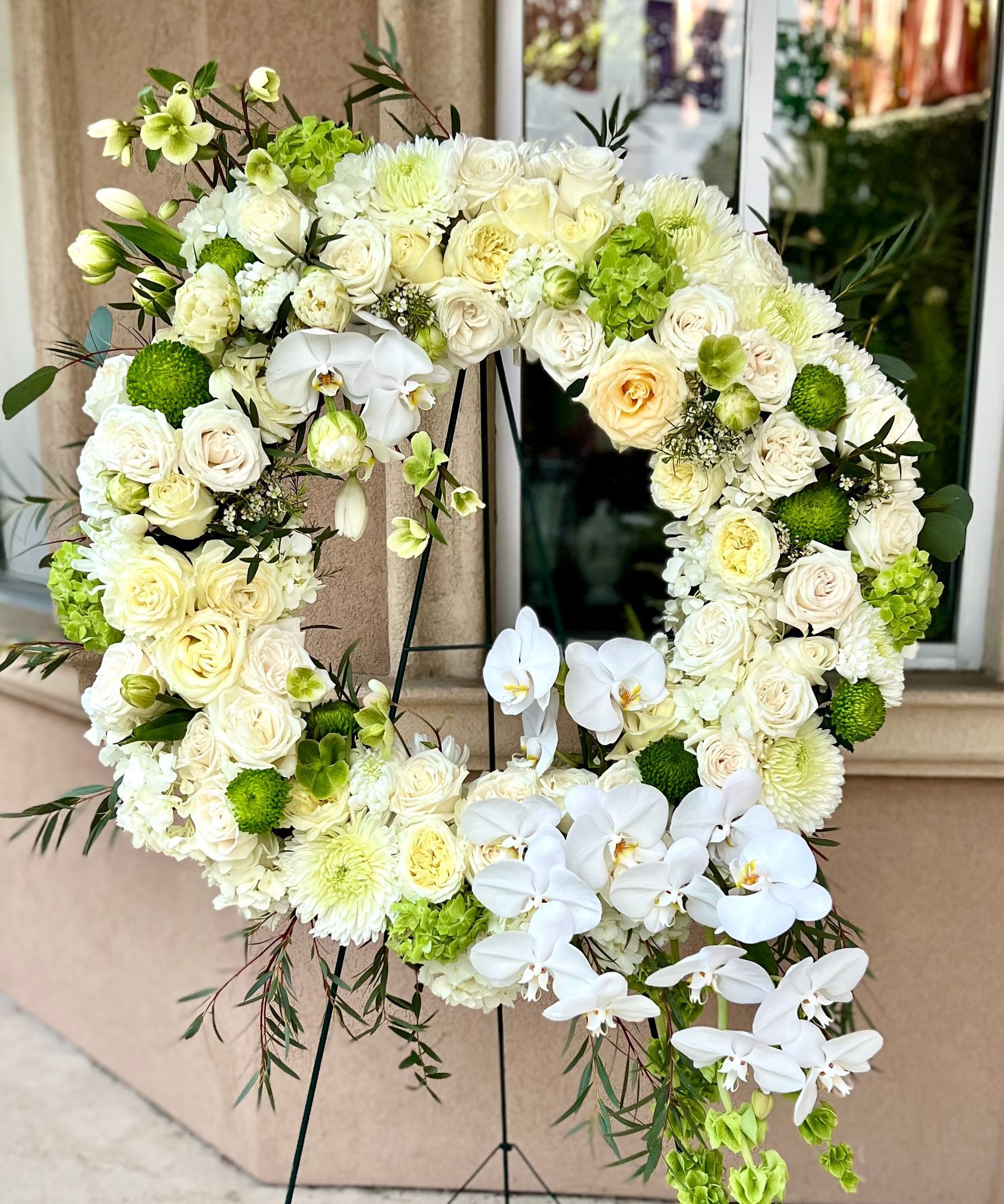 A wreath of white flowers and greenery is sitting on a stand in front of a window.