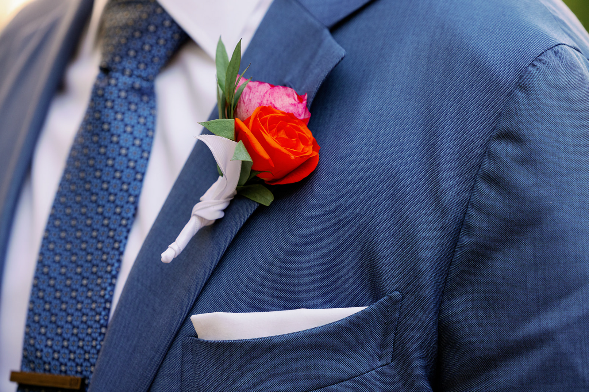 A man in a blue suit and tie has a red rose in his pocket.