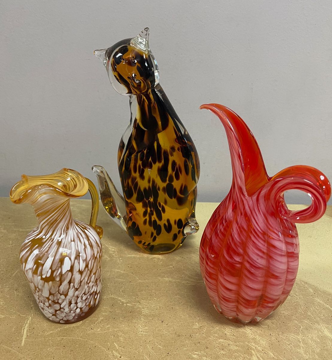 Three glass figurines are sitting on a table including a cat and a vase