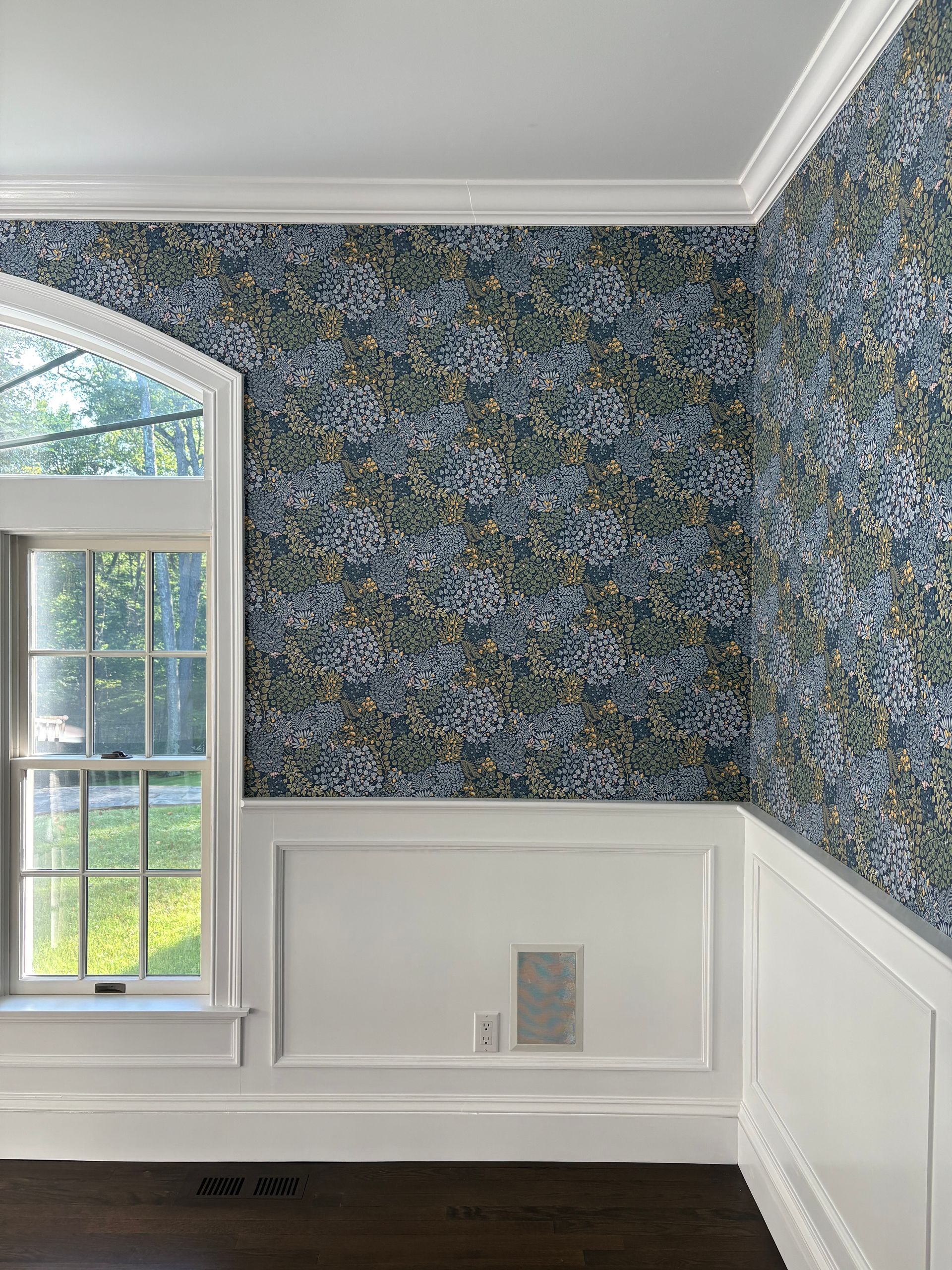 Interior corner with blue floral wallpaper and white wainscoting, featuring a large window and dark wood flooring in a classic dining room.