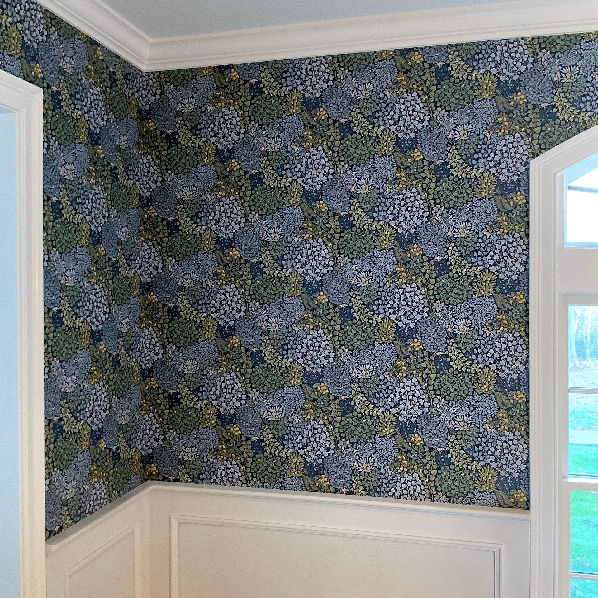 Room corner with dense floral wallpaper, white wainscoting, and natural light from window and doorway, blending ornate and classic elements.