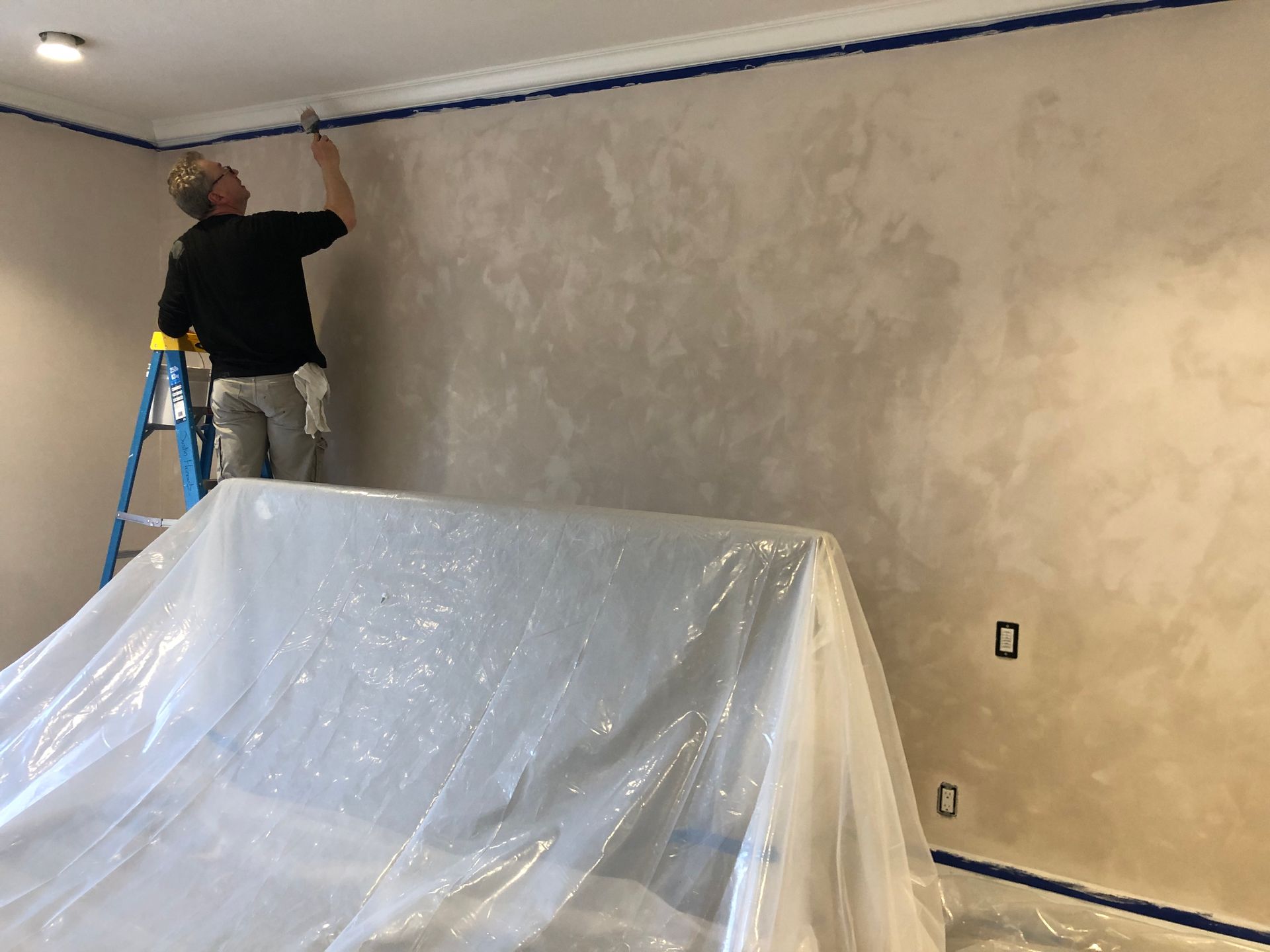 Person on ladder painting upper wall edge near ceiling, with painter’s tape and plastic-covered furniture indicating active home renovation.