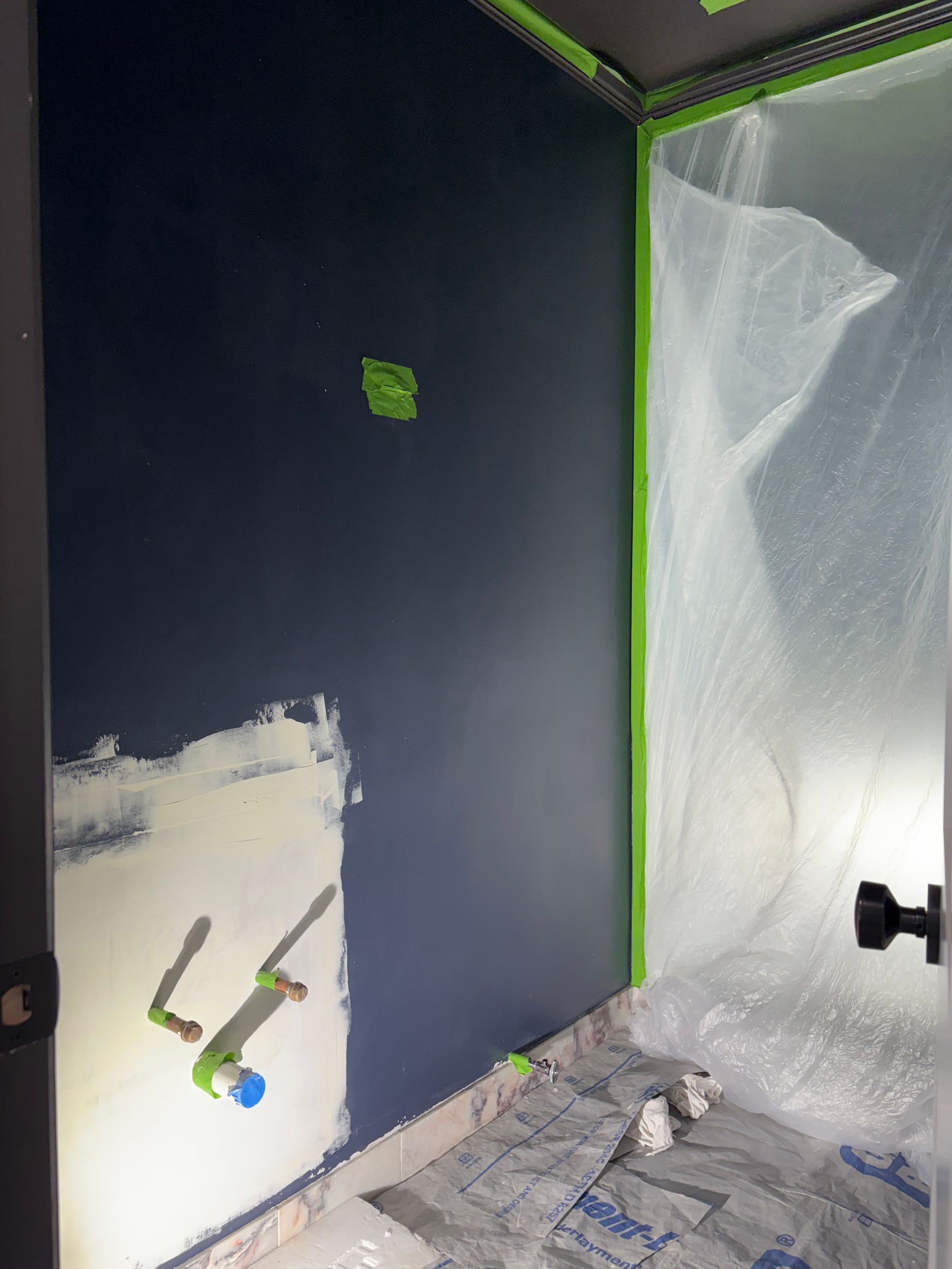 Navy wall with white base coat and exposed plumbing pipes, surrounded by painter’s tape and plastic sheeting during renovation.