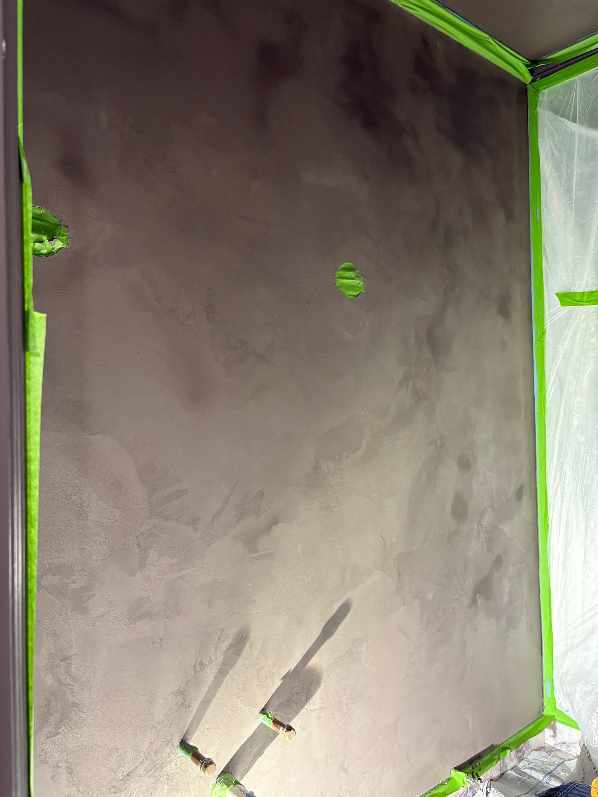 Dark textured wall mid-renovation with green painter’s tape, plastic sheeting, and paint rollers indicating active finishing work.