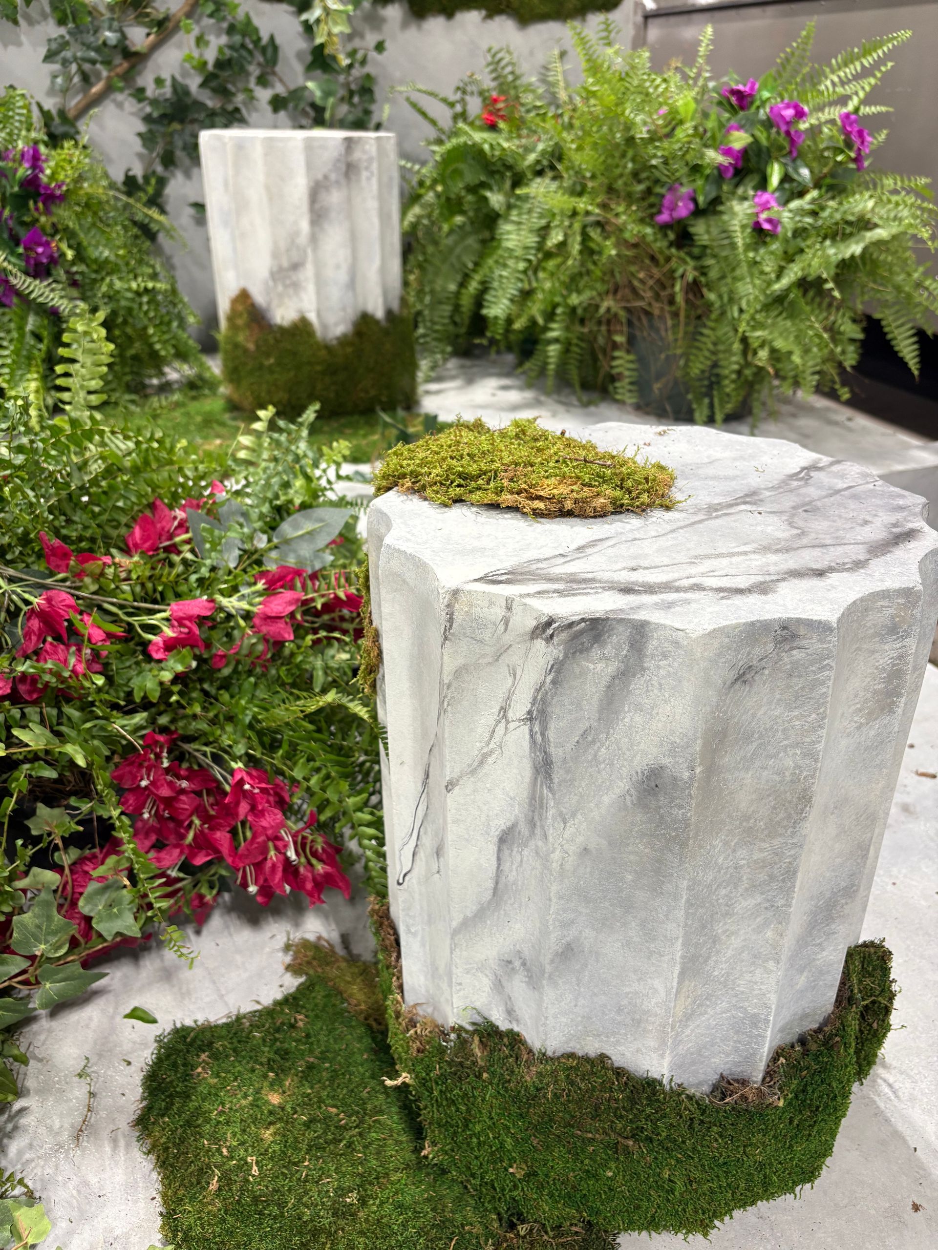 Two fluted marble-like columns with moss accents, surrounded by ferns and colorful flowers.