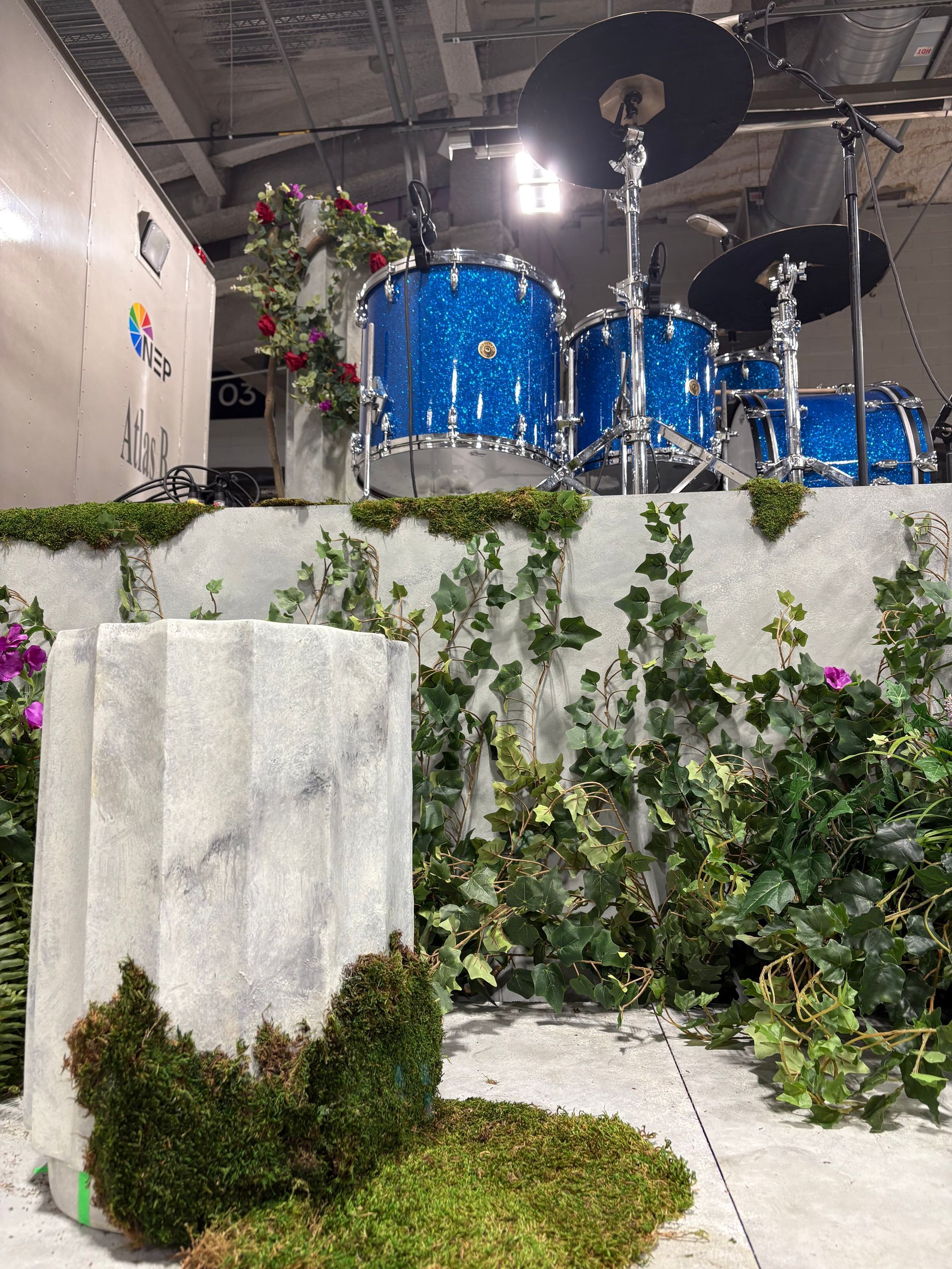 Blue glitter drum kit on a moss-covered platform with faux stone column, blending natural decor with industrial stage elements.