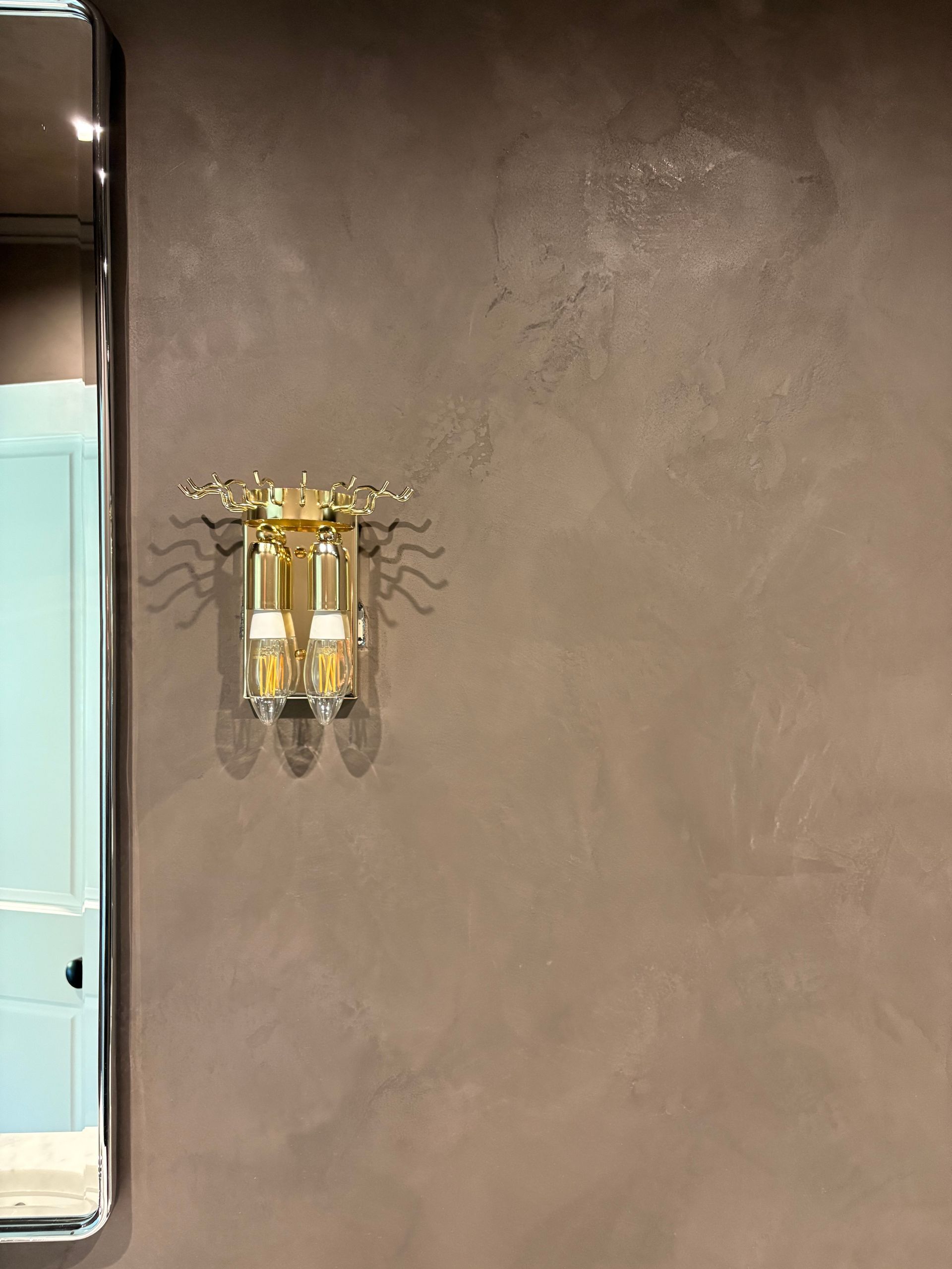 Gold wall light with curved metal arms casting shadows beside a metallic-framed mirror reflecting blue wall and white trim.