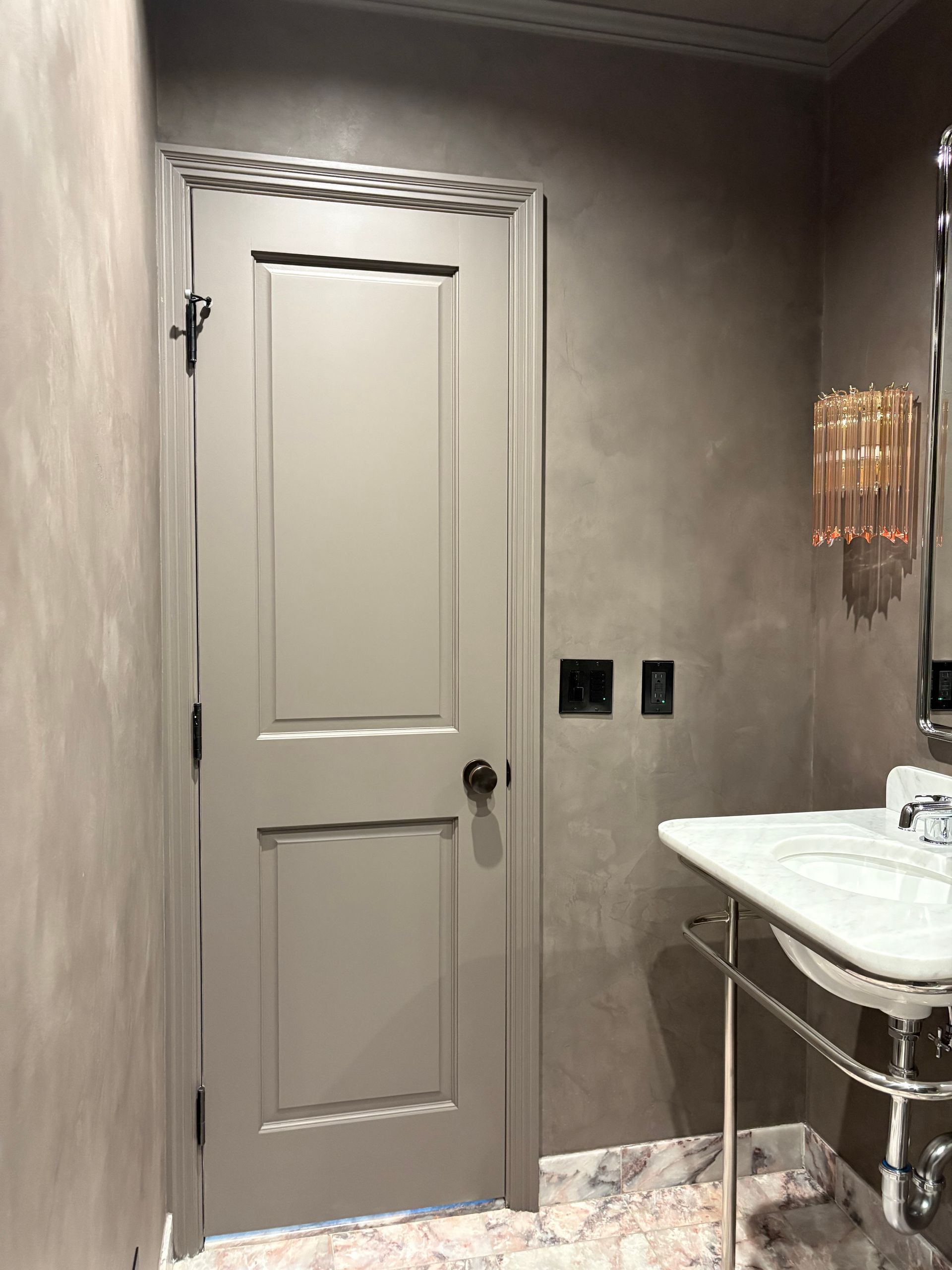 Contemporary bathroom with taupe matte walls, marble floor, chrome-framed sink, and gold vertical light fixture beside black switches.
