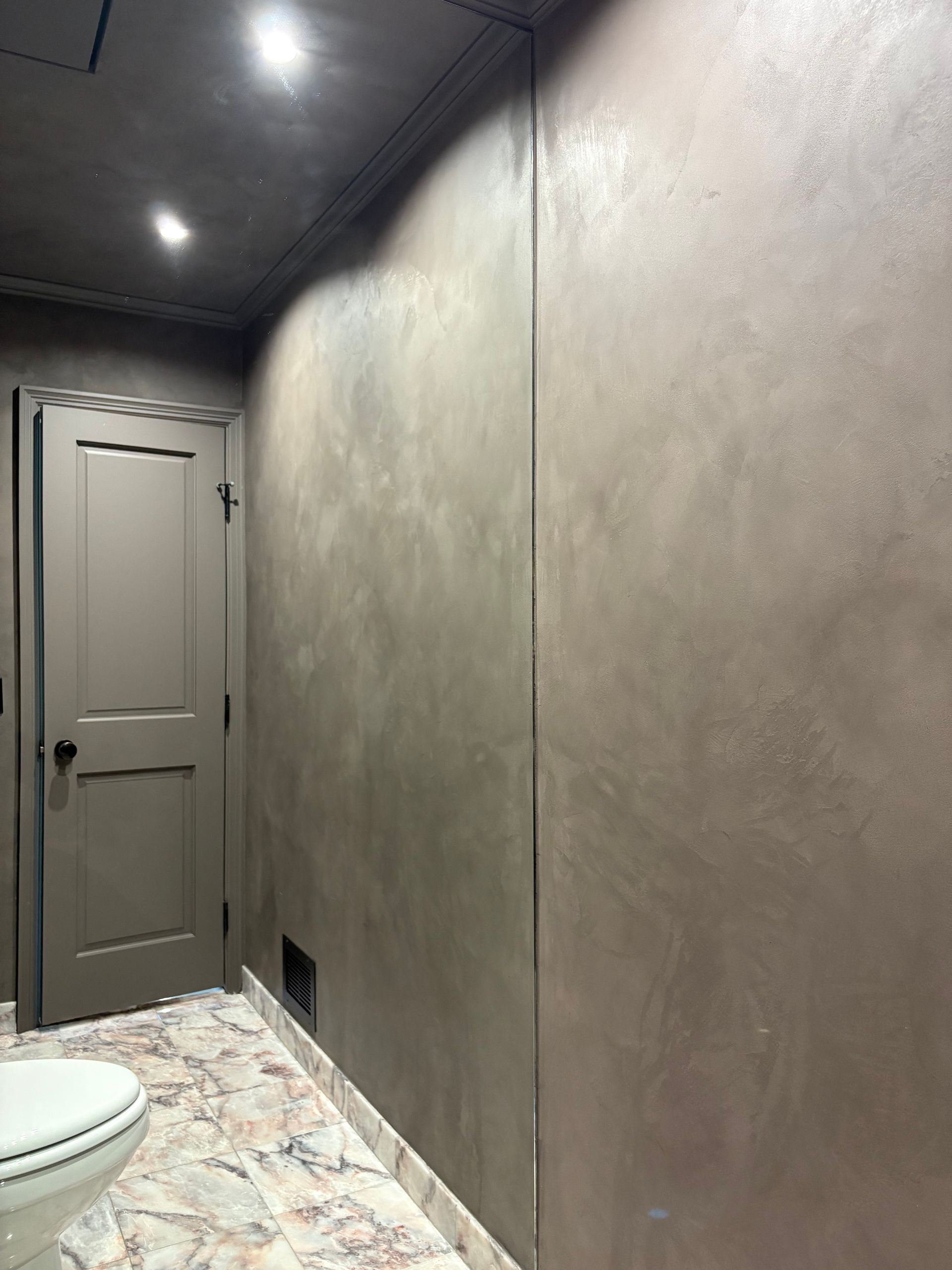Minimalist bathroom with smooth taupe wall, marble tile floor, recessed lighting, and partially visible toilet near paneled door.