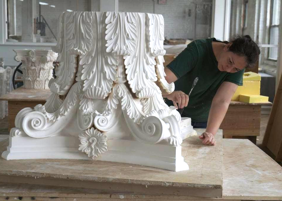 Person working on an ornate white column base with carved acanthus leaves and scrolls in a workshop setting.