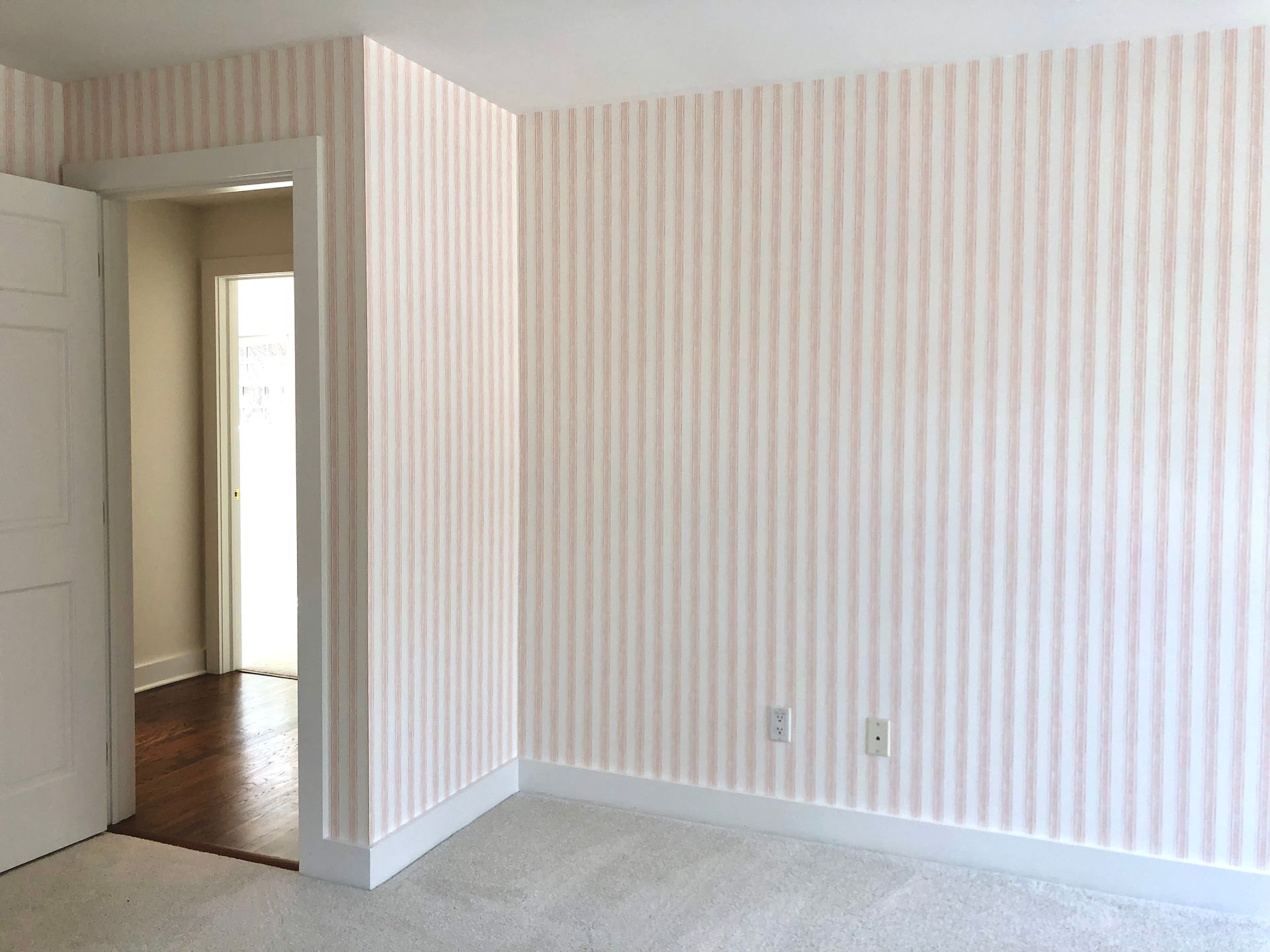 Interior corner with light pink and white striped wallpaper, white baseboards, and doorway leading to a room with wood flooring.