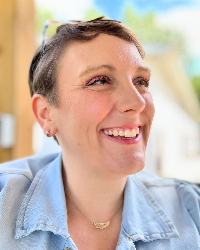 A person with short hair smiles warmly, looking off to the side, wearing a light blue denim shirt and a gold necklace.
