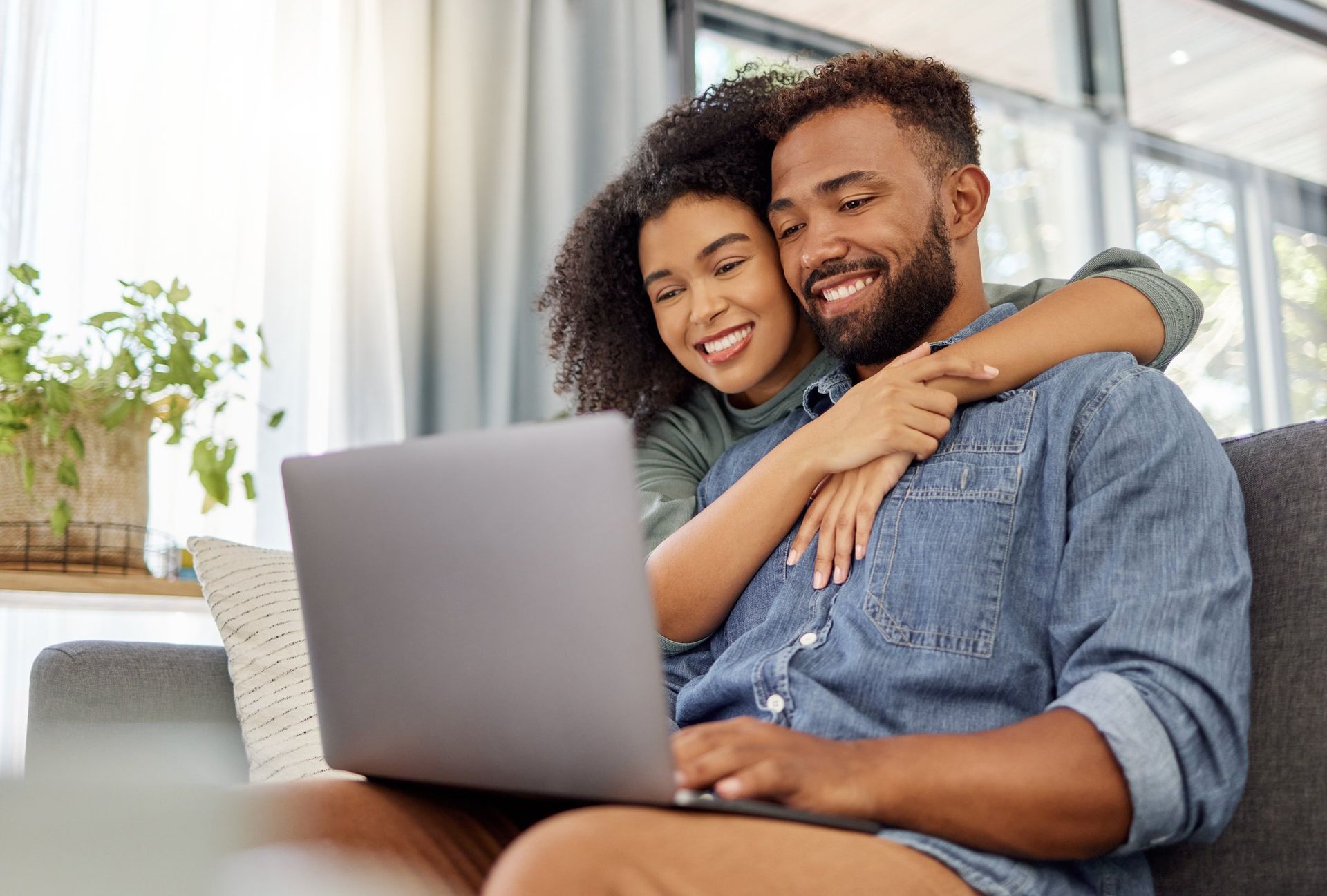 A couple sits together on a sofa, smiling while looking at a laptop computer.