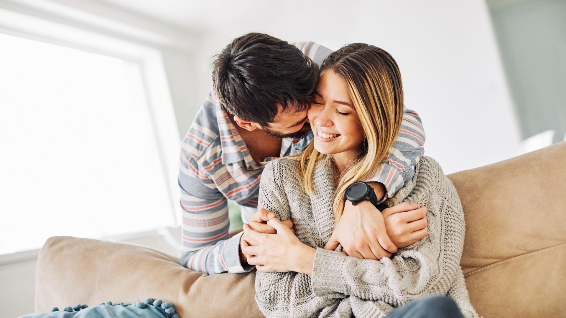 A person in a plaid shirt embraces another person in a chunky grey knit sweater from behind while whispering in their ear.