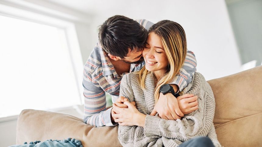 A person in a plaid shirt embraces another person in a chunky grey knit sweater from behind while whispering in their ear.