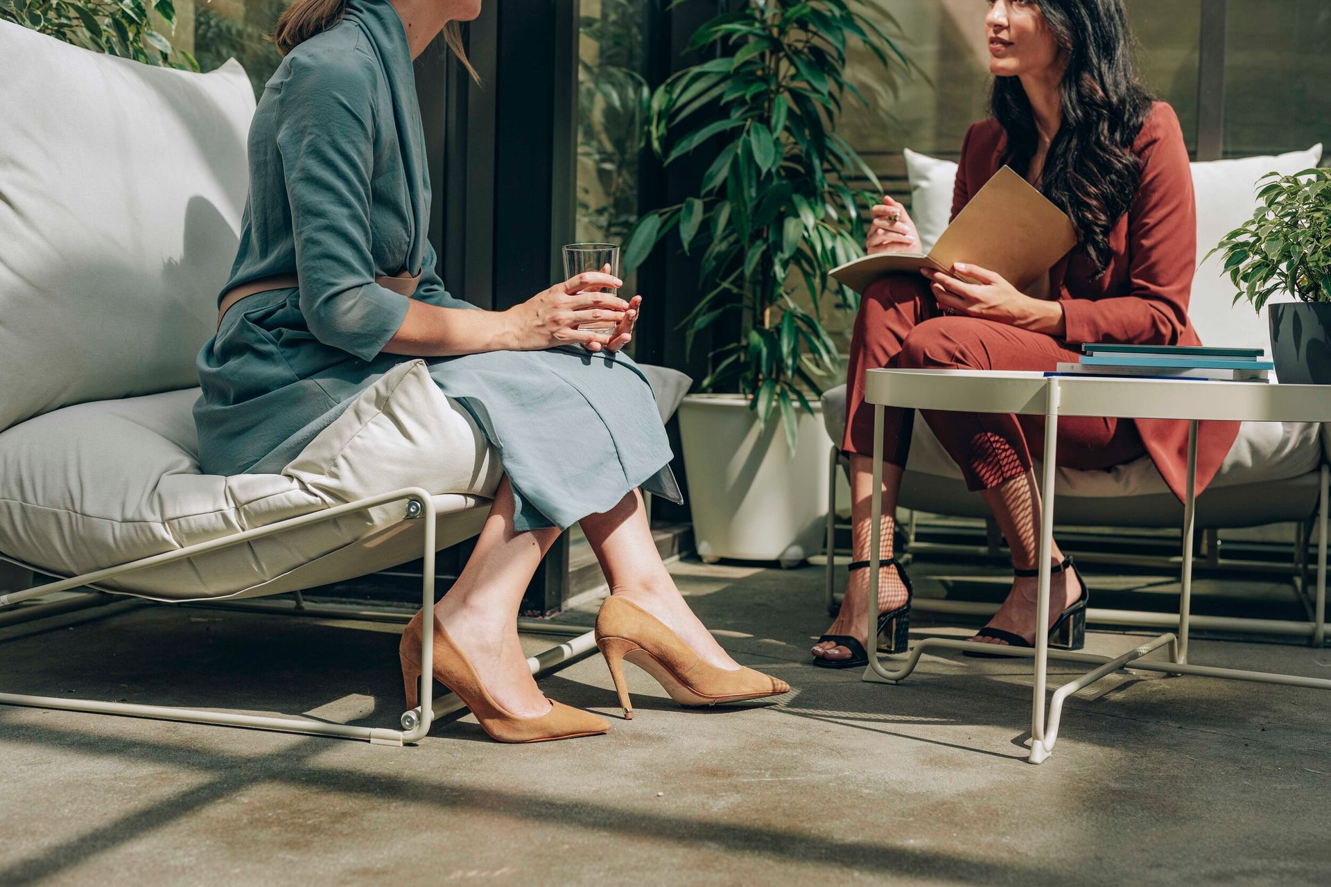 Two individuals sit in chairs on a patio, one in a blue dress and one in a rust suit, discussing notes over a table.