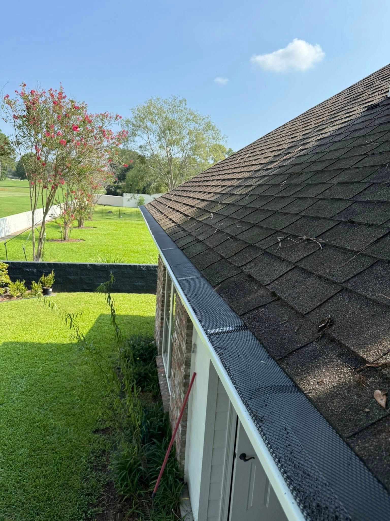 Gutter with black mesh cover on a roof, view of the side of a house with green grass and trees.