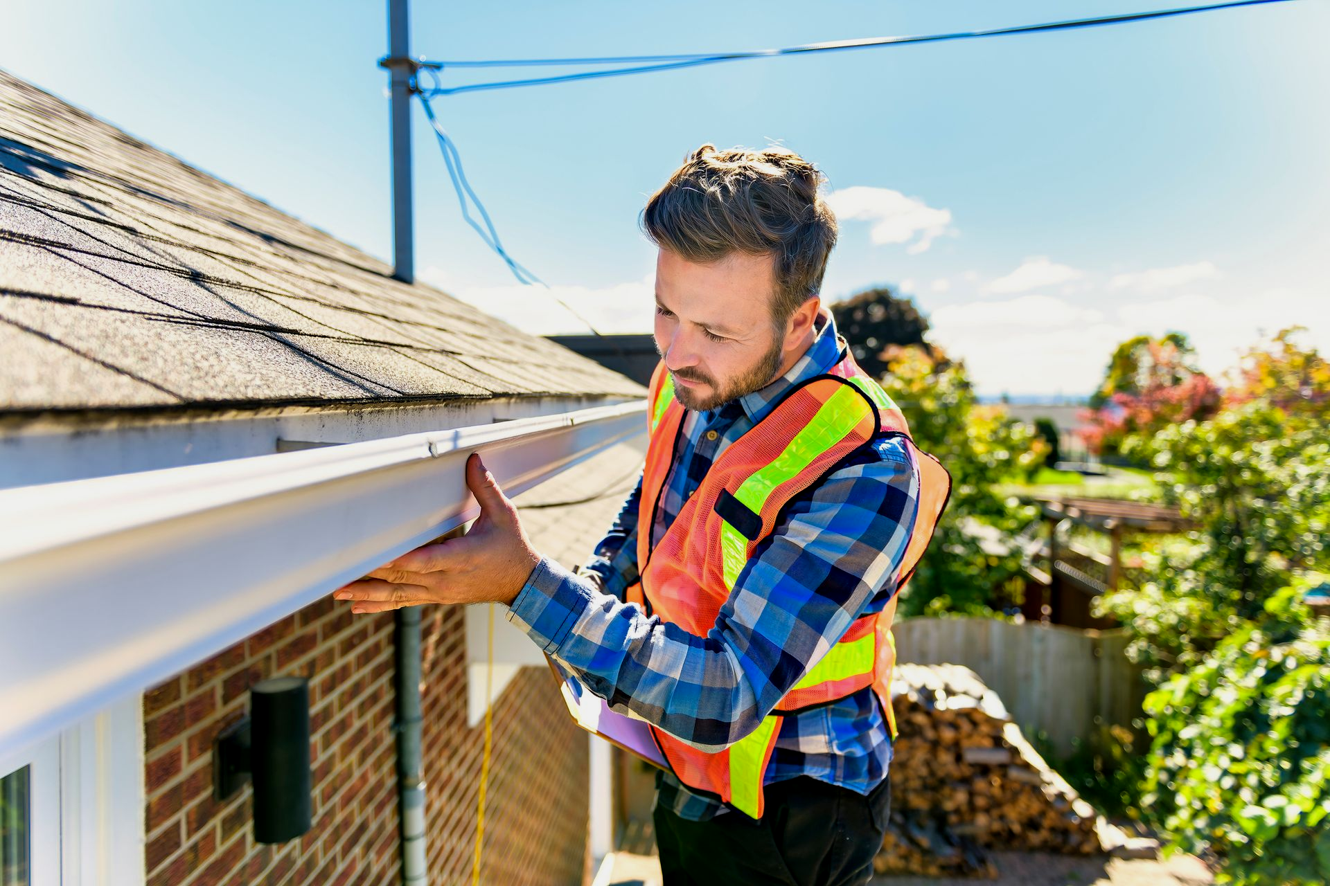 Man in a safety vest installing a white gutter on a brick house roof. Sunny day.
