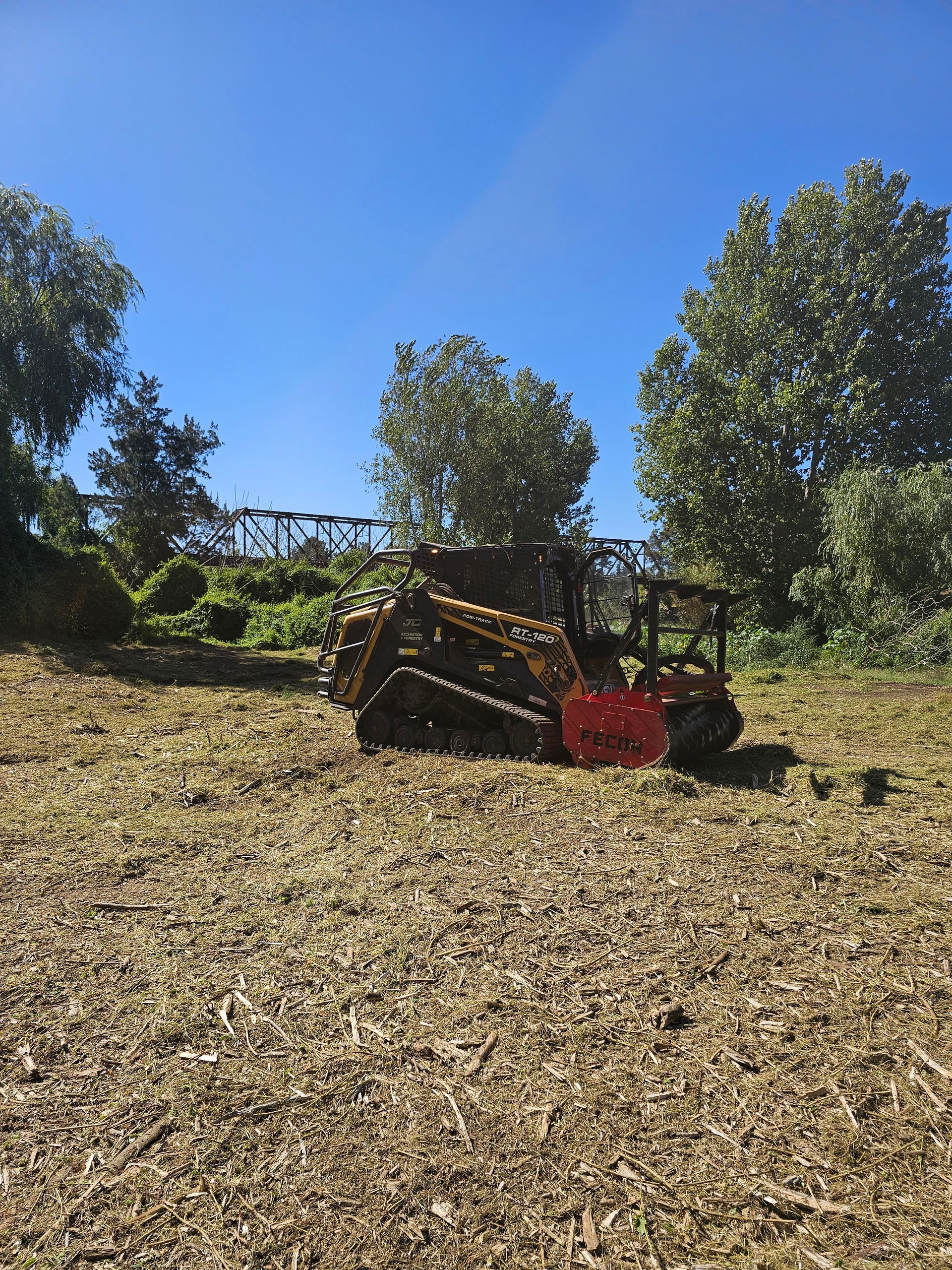A yellow tracked skid steer with a forestry mulcher attachment working in a cleared field under a clear blue sky.