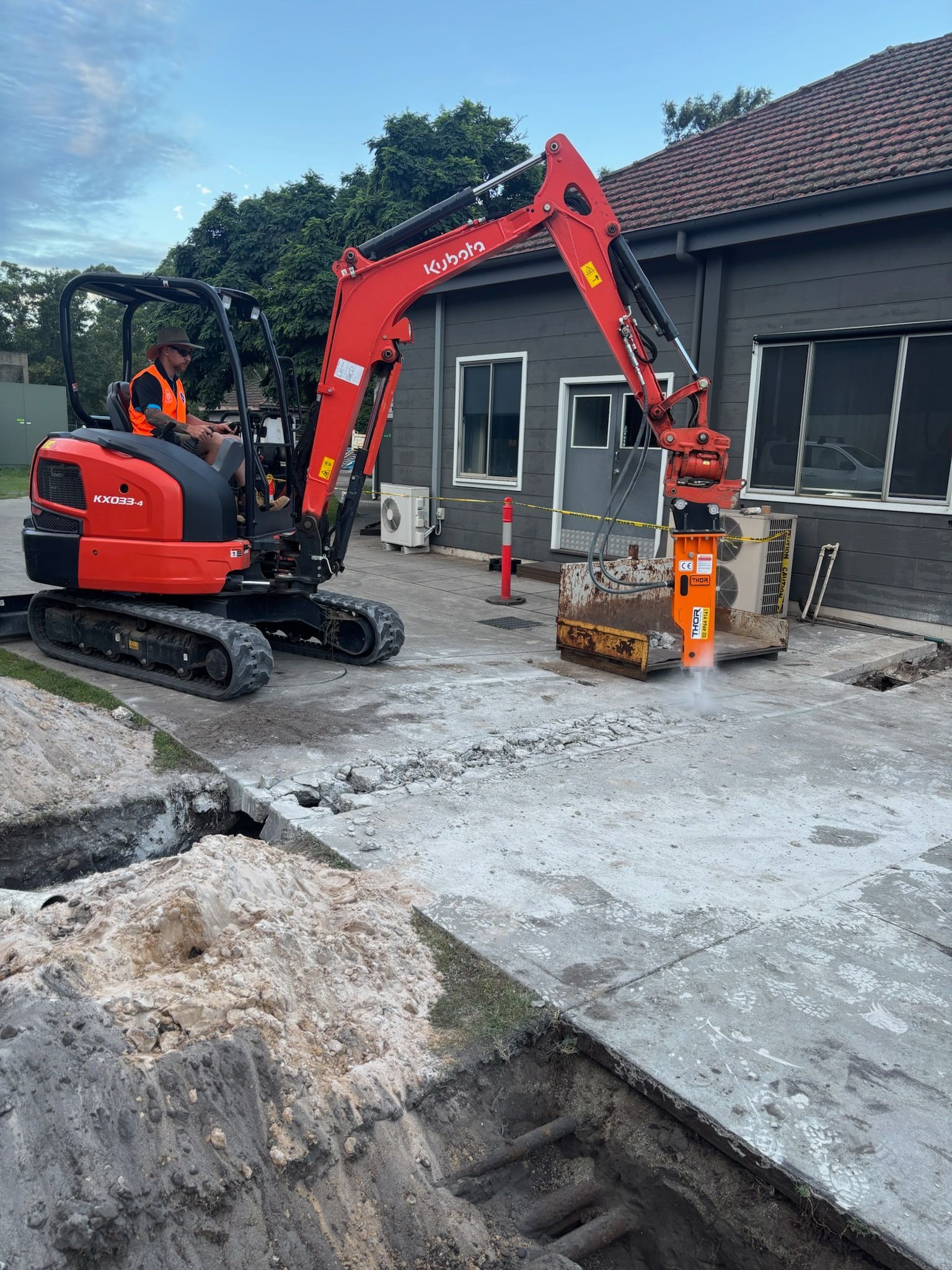 Red excavator demolishing concrete near a building with a worker inside— JC Excavation and Mechanical Pty Ltd in Raymond Terrace, NSW