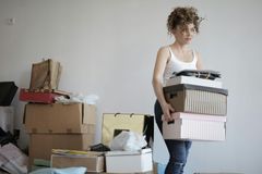 A woman with curly hair in a tee shirt carrying several boxes, with stacks of boxes behind her.