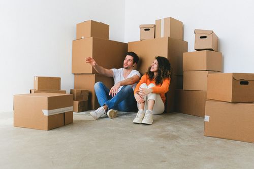 A couple sitting on the floor with a lot of packed cardboard boxes. They are looking up towards the top of the room.