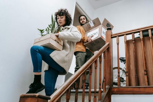 A couple in sweaters and warm clothing walking down wooden stairs carrying boxes. 