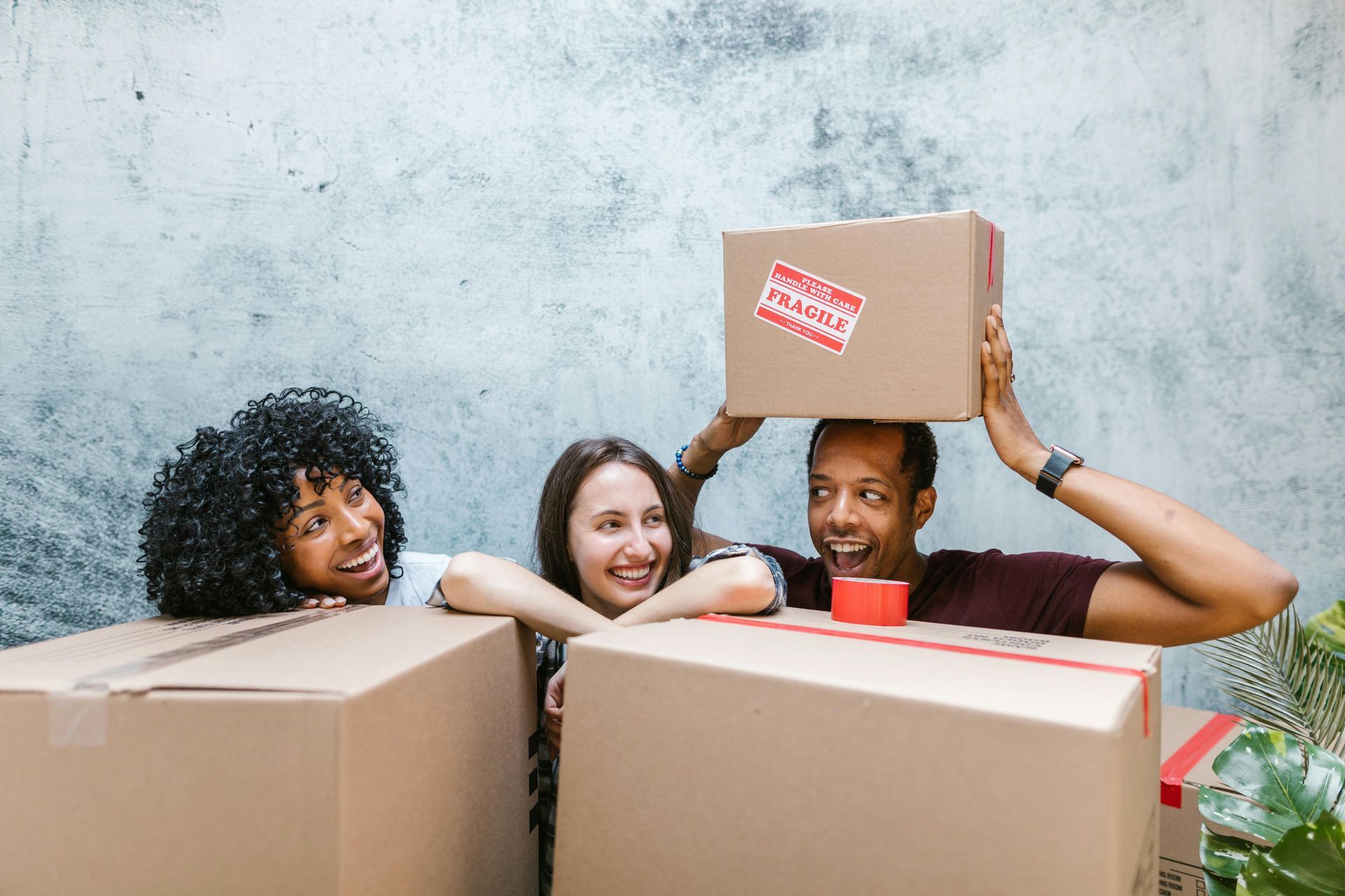 A group of two women and a man near moving boxes. The women are looking at the man who is holding a fragile box on top of his head in a funny manner.