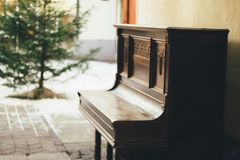 An old dark brown wooden piano with the key cover closed, outside on a patio. There is a small pine tree in the middle of the patio area.