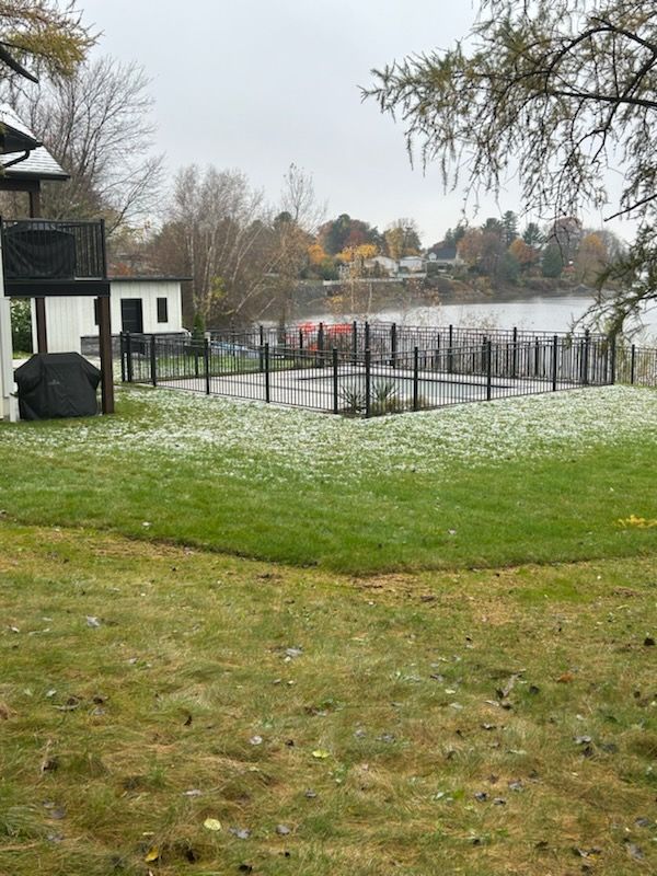 Une pelouse enneigée mène à une piscine clôturée en noir au bord d'un lac, avec des maisons sur la rive lointaine.
