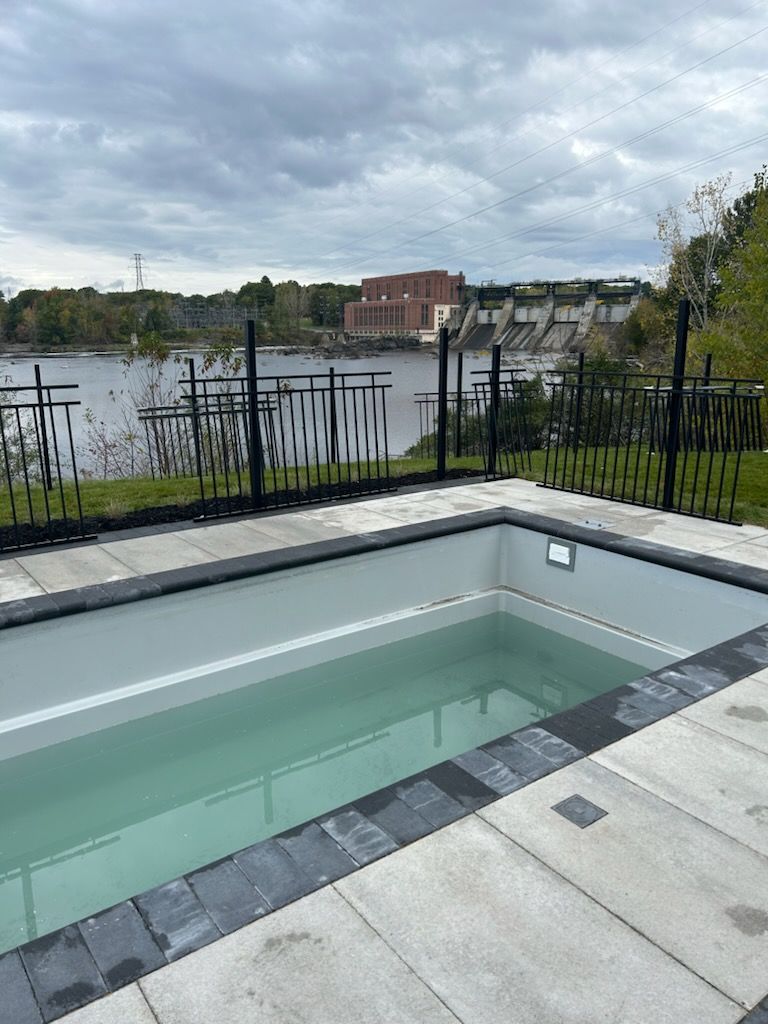 Piscine rectangulaire extérieure avec eau, clôture noire, rivière avec barrage en arrière-plan, ciel nuageux.