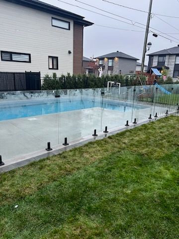 Jardin avec piscine entourée d'une clôture en verre, d'herbe verte et de maisons. Ciel couvert.