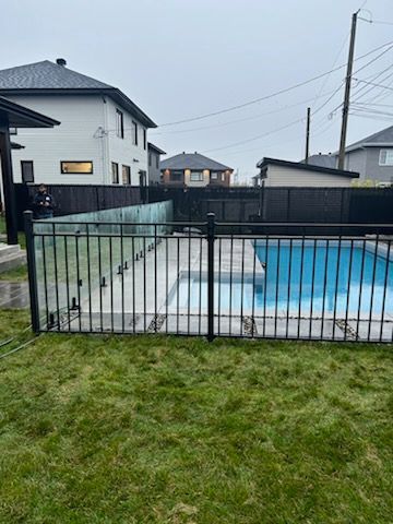 Piscine clôturée noire entourée d'herbe verte, maisons en arrière-plan par une journée nuageuse.