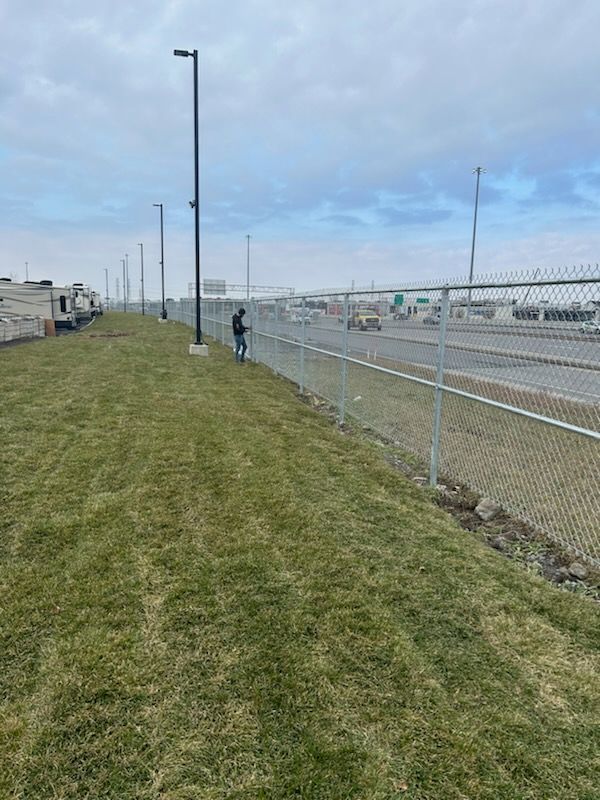Une personne se tient près d'une clôture grillagée bordant une zone herbeuse. La route et le ciel couvert sont visibles en arrière-plan.
