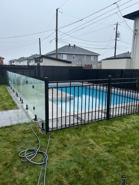 Clôture de piscine en métal noir et en verre entourant une piscine dans le jardin par une journée nuageuse.