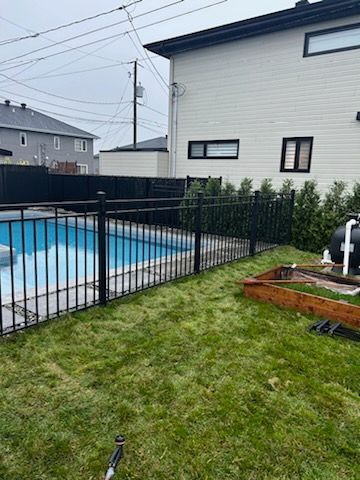 Une clôture noire entoure une piscine dans l'arrière-cour et de l'herbe verte, une maison blanche en arrière-plan.