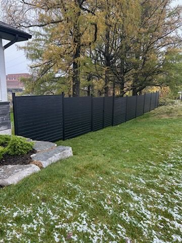 Clôture à lattes horizontales noires le long d'une pelouse herbeuse avec des arbres en arrière-plan.