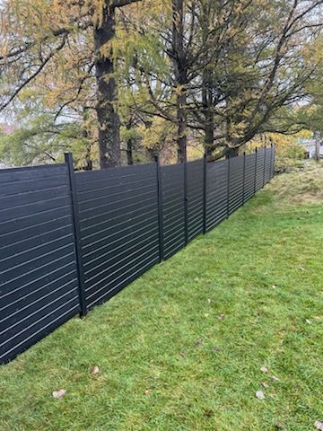 Clôture à lattes horizontales noires le long de l'herbe verte ; arbres en arrière-plan.