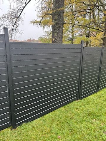 Clôture à lattes horizontales noires dans une cour herbeuse avec un arrière-plan d'arbres.