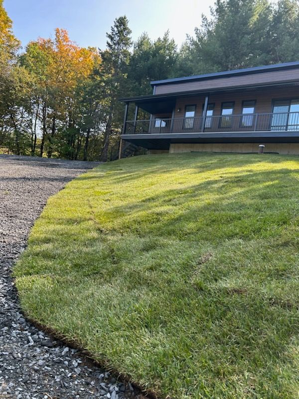Une pelouse verte luxuriante monte jusqu'à un bâtiment marron avec un porche ; une allée de gravier est au premier plan ; des arbres aux couleurs d'automne sont à l'arrière-plan.