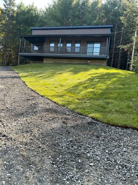 Une cabane moderne avec une allée de gravier et une colline herbeuse, située dans une zone boisée.