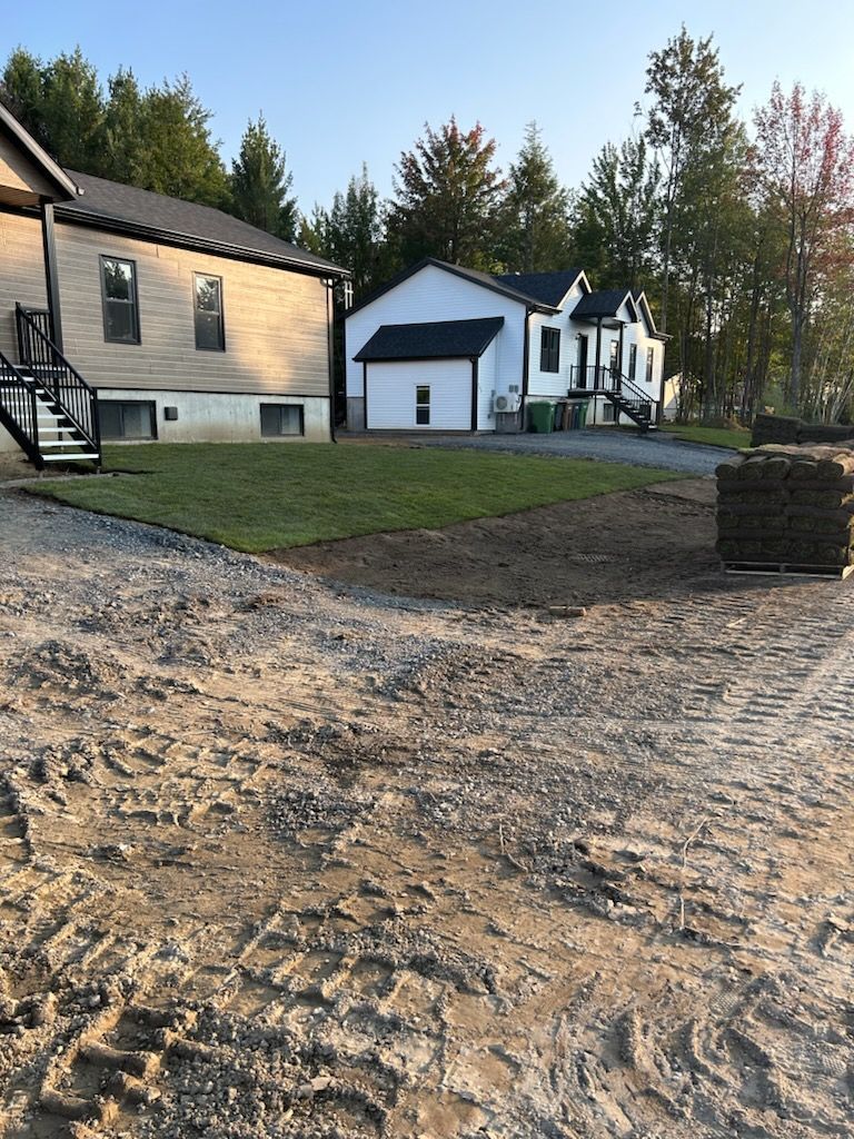 Maisons en construction avec nouveau gazon sur un terrain nivelé, gravier au premier plan, arbres et ciel clair.