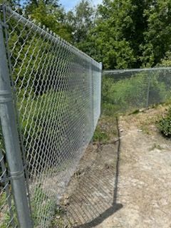 Clôture grillagée dans une zone herbeuse avec une allée en ciment, des arbres en arrière-plan.