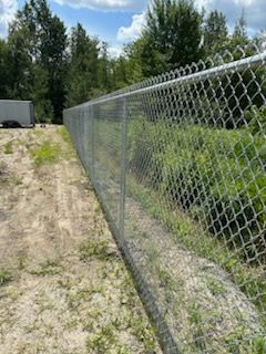 Une clôture grillagée avec du fil barbelé le long de son sommet, dans un champ herbeux avec des arbres en arrière-plan.