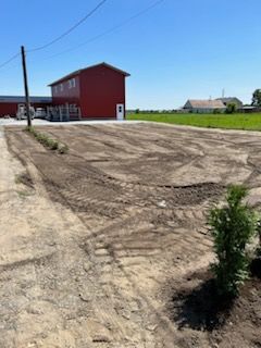 Aire de stationnement en terre devant un bâtiment rouge sous un ciel bleu ; des jeunes arbres sont plantés.
