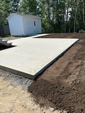 Une fondation en dalle de béton dans une cour, à côté de terre et d'un petit bâtiment blanc en arrière-plan.