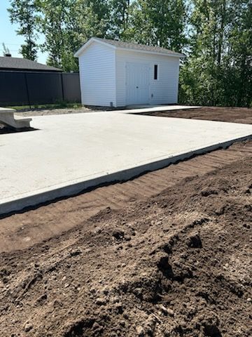 Patio en béton avec un cabanon blanc dans une cour arrière, entouré de terre et d'arbres verts.