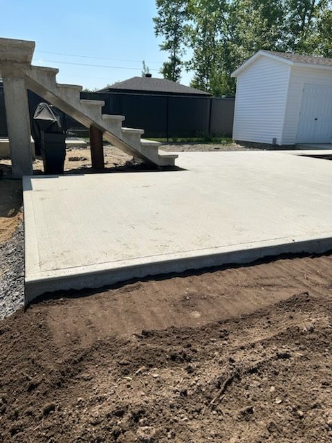 Patio en béton avec escaliers, sol foncé au premier plan, un garage blanc et une clôture en arrière-plan.